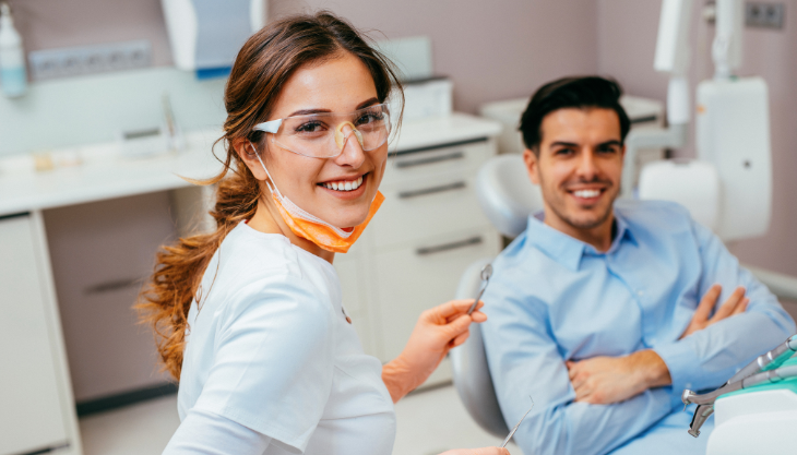 A female dentist holding up a concave mirror with a patient sitting on the dental chair in the background.