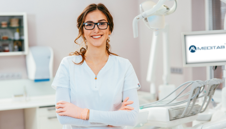A female dental professional smiling at the camera crossing her arms with a dental chair in the background.