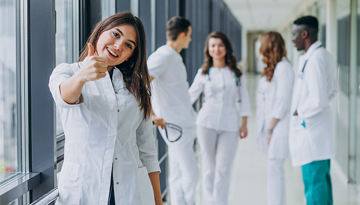 A female doctor gesturing the thumbs up while 4 other healthcare workers huddle in the background.