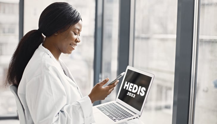 A doctor holding a phone and a laptop with 2023 HEDIS measures showing on screen.