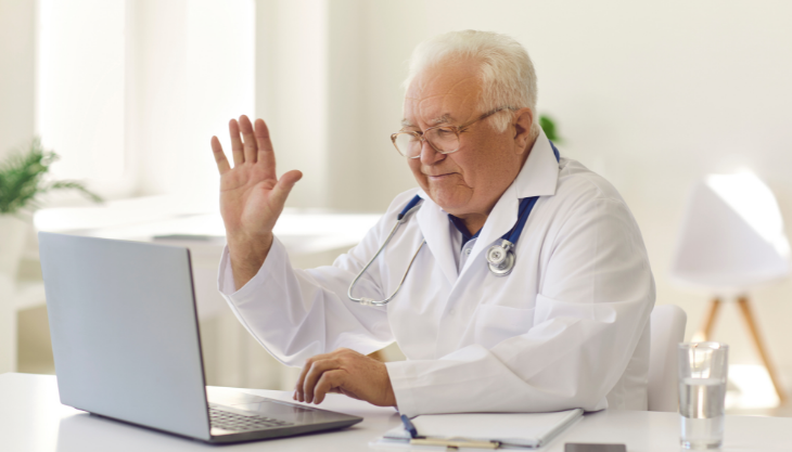 Doctor waving at a laptop screen during a telehealth video call.